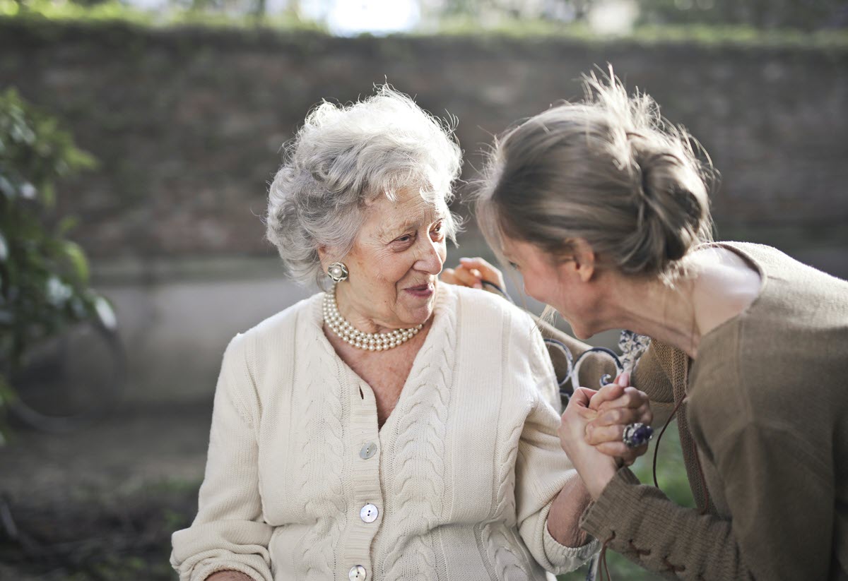 Elderly woman smiling and holding hands with a younger woman outdoors.